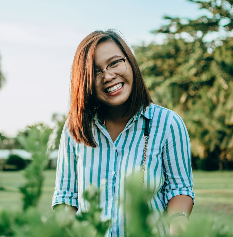 Smiling woman wearing glasses and a blue-and-white striped shirt standing outdoors in a green park on a sunny day.