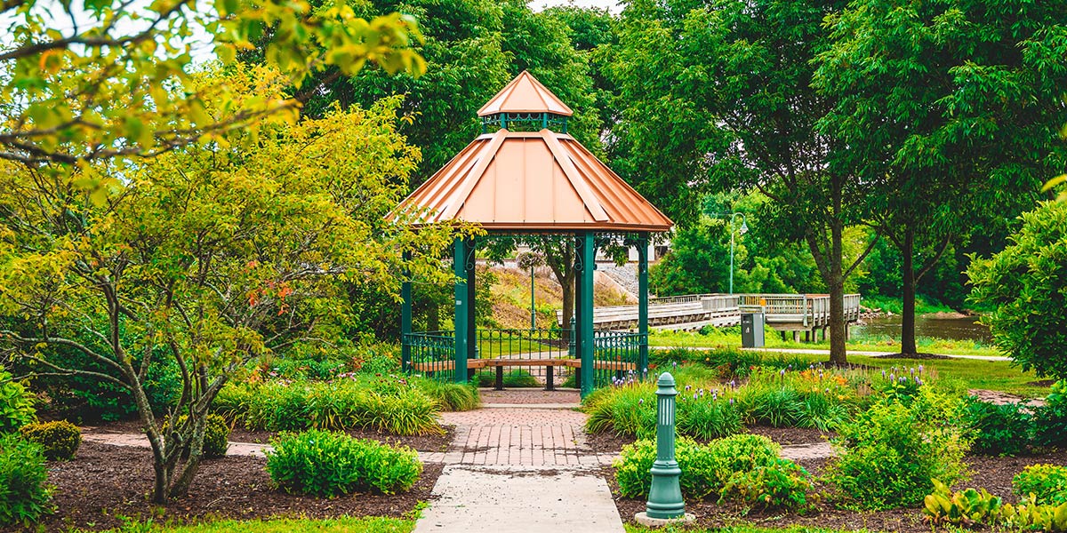 A small copper-roofed gazebo in a vibrant garden, surrounded by trees, flowers, and a river boardwalk.